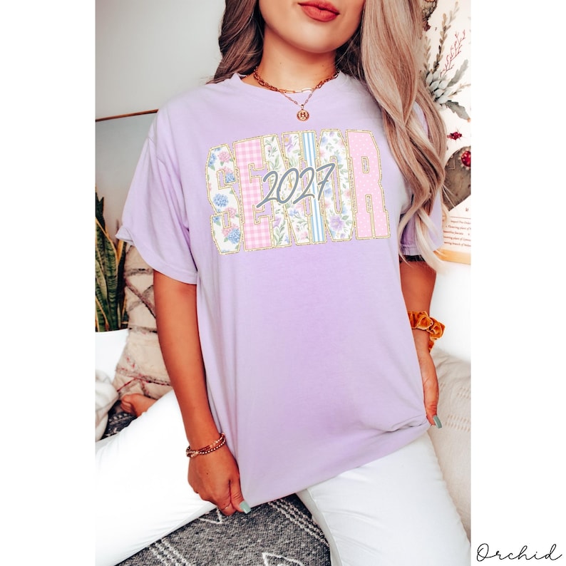 May include: A light purple short-sleeve t-shirt featuring the word SENIOR in large block letters filled with a mix of floral, gingham, and striped patterns in pastel colors. The number 2027 is printed in a dark gray script font over the center.