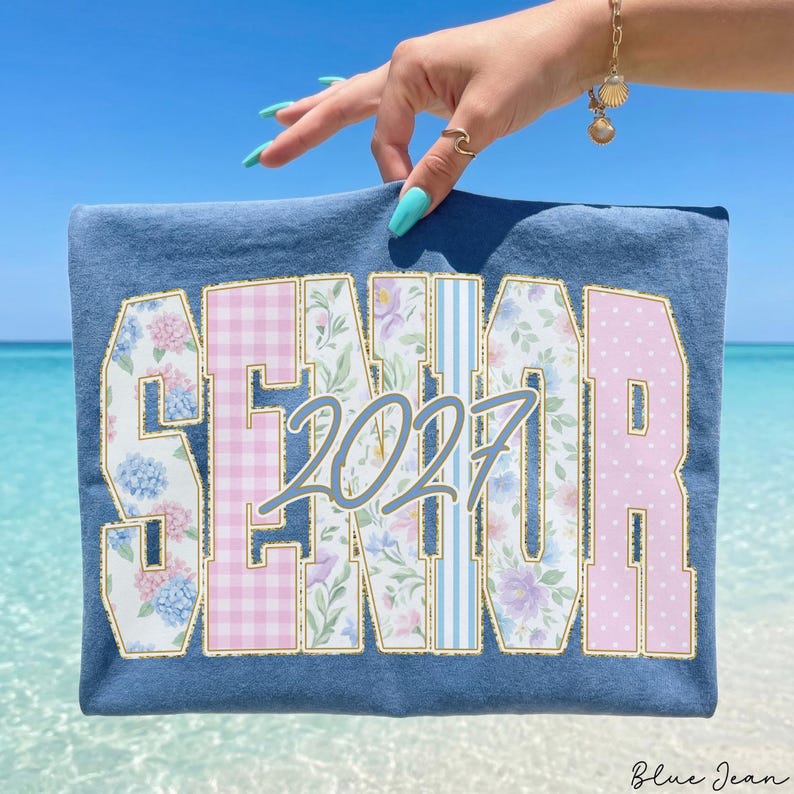 May include: A blue t-shirt featuring the word SENIOR in large block letters with varied patterns including floral prints, pink gingham, and blue stripes. The number 2027 is printed in a cursive font over the center of the word SENIOR.