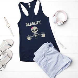 May include: Navy blue tank top with the word "DEADLIFT" and a skeleton lifting weights graphic. The image also includes a pair of silver sandals, a watch, light blue shorts, and pink headphones.