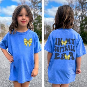 K&ouml;nnte beinhalten: Ein blaues T-Shirt in Kindergr&ouml;&szlig;e mit einer gelben Softball-Schleife auf der Vorderseite und dem Text "In My Softball Era" mit einer gelben Softball-Schleife auf der R&uuml;ckseite.