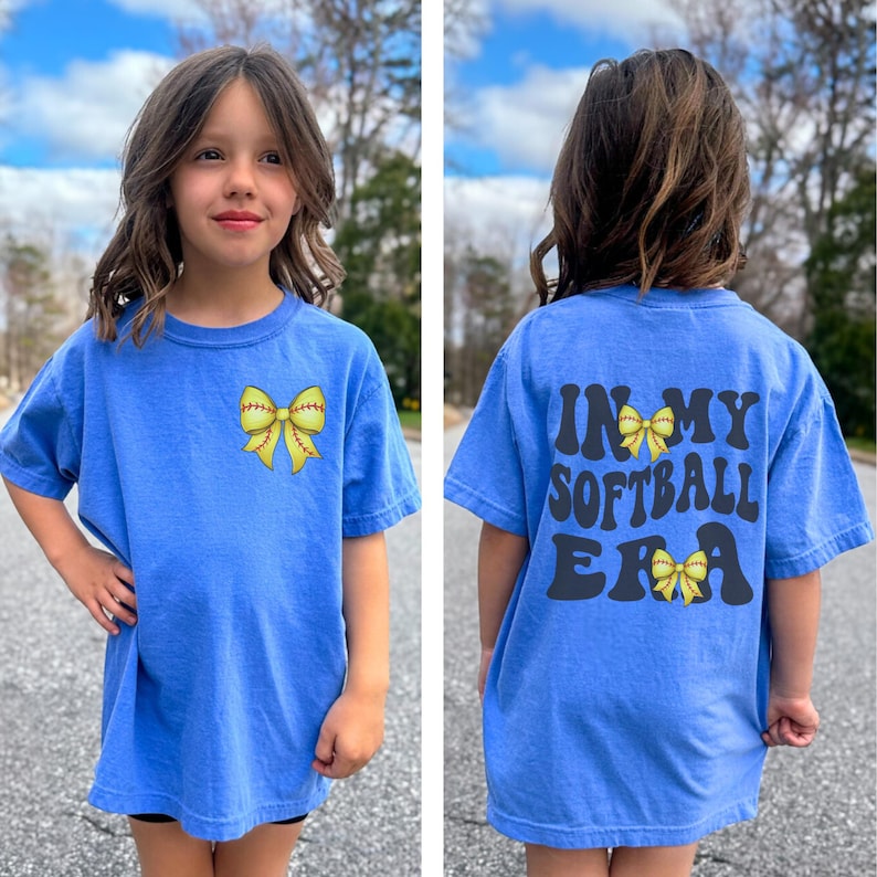 K&ouml;nnte beinhalten: Ein blaues T-Shirt in Kindergr&ouml;&szlig;e mit einer gelben Softball-Schleife auf der Vorderseite und dem Text "In My Softball Era" mit einer gelben Softball-Schleife auf der R&uuml;ckseite.