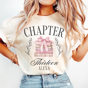 May include: A white t-shirt with the text "CHAPTER THIRTEEN ALEXA" and a watercolor illustration of a stack of books tied with a pink ribbon.
