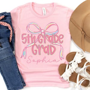 May include: A pink t-shirt with the text "5th Grade Grad Sophia" in a colorful bow design. The image also includes blue jeans, a pink polka dot hair tie, a tan sun hat, and white sandals.