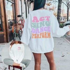 May include: White sweatshirt with the words "ACT SING DANCE PERFORM" in colorful retro font on the back. The front of the sweatshirt has a small graphic of two theater masks with the word "Camilla" underneath.