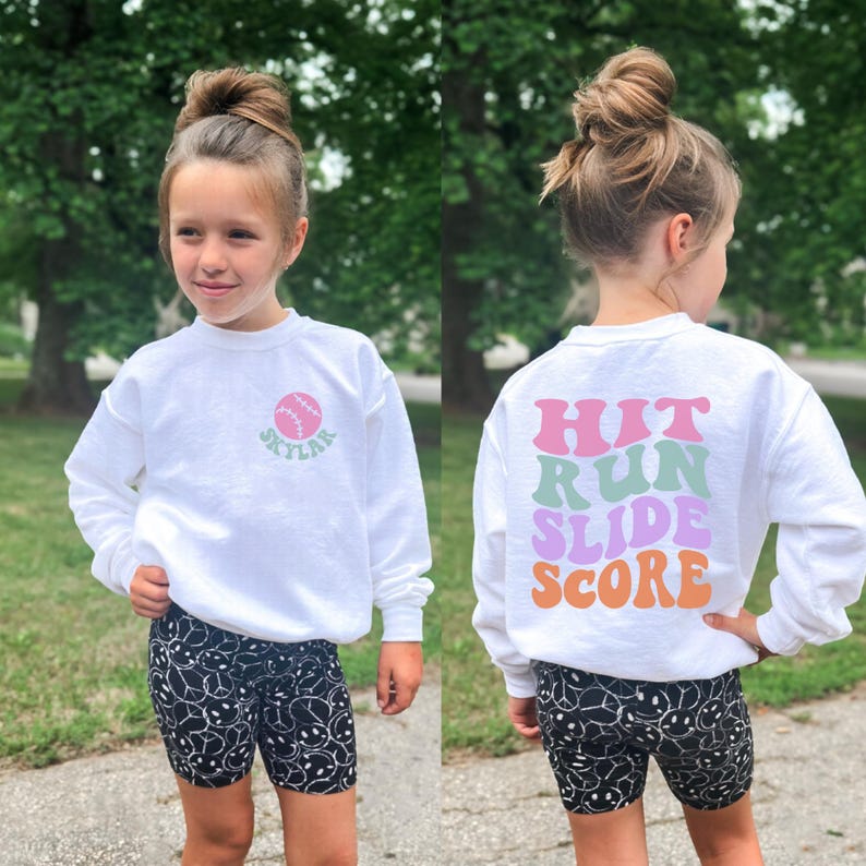 May include: A young person wearing a white sweatshirt with a baseball graphic and the name "Skylar" in green. The back of the sweatshirt has the words "Hit Run Slide Score" in various colors. They are wearing black shorts with a white smiley face pattern.