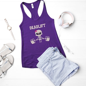 May include: A purple racerback tank top with the word "DEADLIFT" above a skeleton lifting weights. The image also includes a pair of silver sandals, a watch, headphones, and denim shorts.