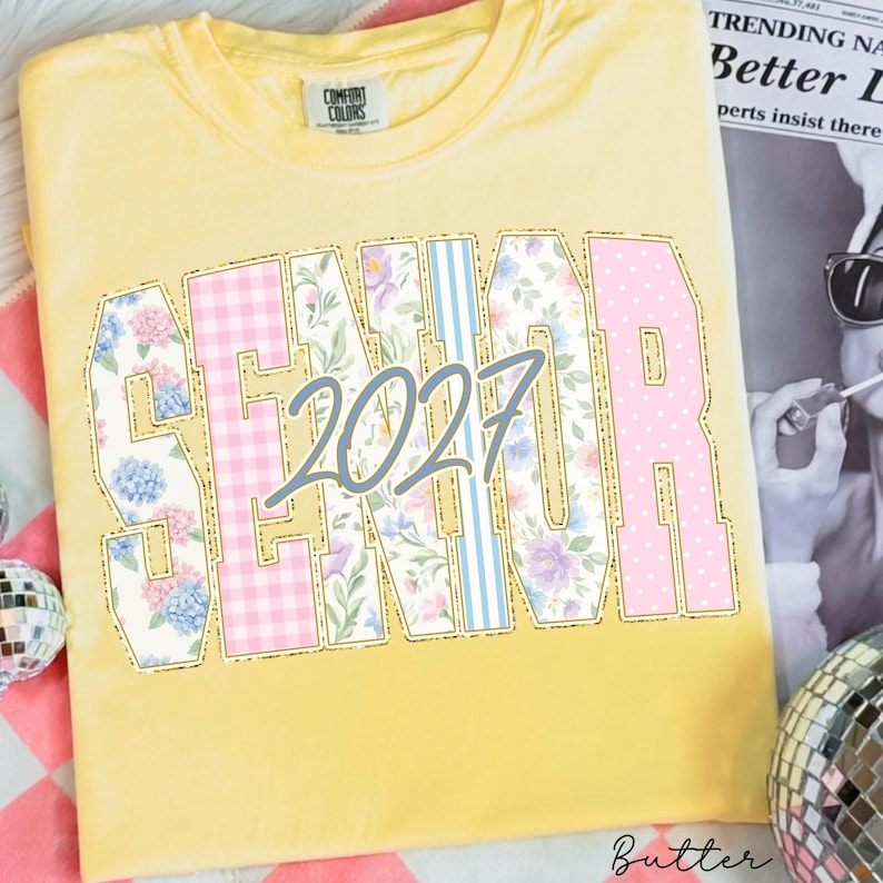 May include: A yellow short-sleeved t-shirt featuring the word SENIOR in large block letters with varied patterns including pink gingham, floral prints, and blue stripes. The number 2027 is printed in a grey script font across the center.
