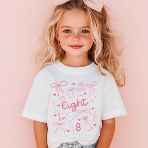 Op de afbeelding: Wit T-shirt met roze verjaardagsthema graphics en de tekst "Eight" op de voorkant.