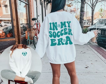 Może przedstawiać: Biała bluza z zielonym nadrukiem, na którym widnieje napis "In My Soccer Era" z piłką nożną w tekście. Bluza ma zielone serce z piłką nożną w środku z przodu.
