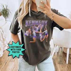 May include: A dark gray t-shirt with the word "KENNEDY" printed in purple letters. The shirt features a photo of a group of young women performing gymnastics. The text "CUSTOMIZE WITH YOUR PHOTOS" is in a teal starburst.
