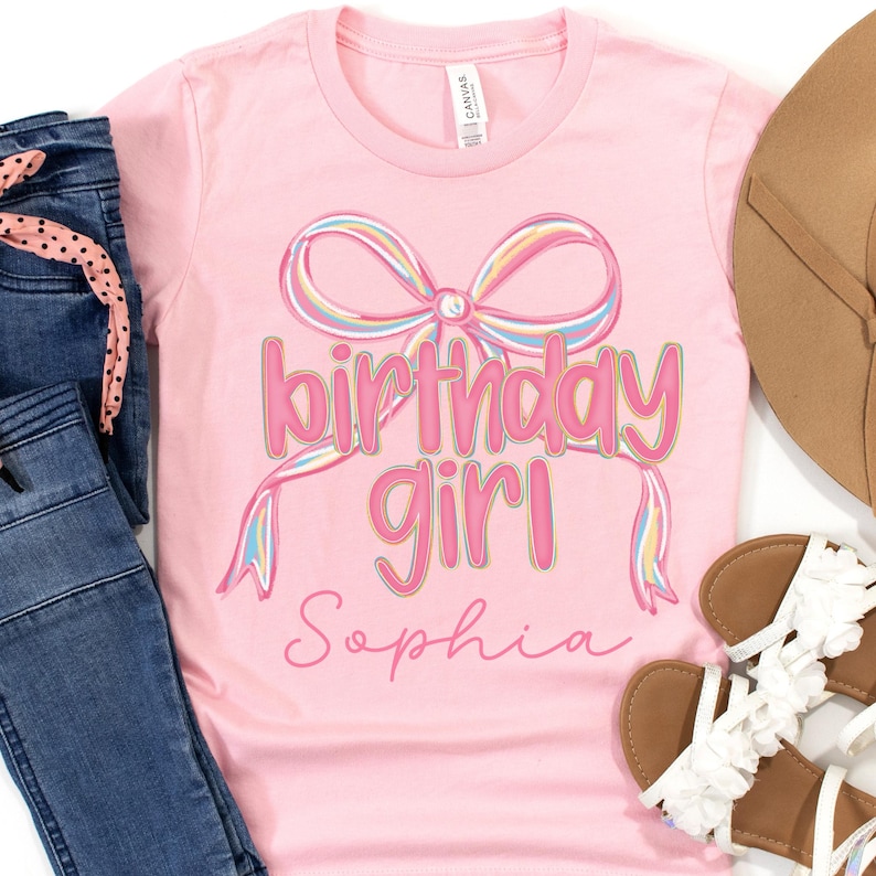 以下が含まれることがあります： ピンク色のTシャツ。ピンク色の「birthday girl」の文字とパステルレインボーのリボンが描かれています。「Sophia」という名前が筆記体で下に書かれています。青いジーンズと白いサンダルと一緒に表示されています。