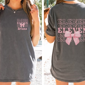 May include: A gray t-shirt with a pink bow and the word "ELEVEN" repeated multiple times. The shirt also has the name "Briana" printed on the front.