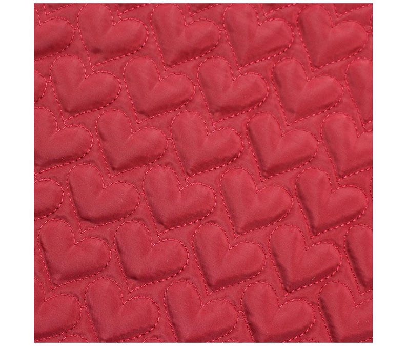 May include: Close-up of a red fabric with a quilted heart pattern. The hearts are evenly spaced and stitched with a contrasting thread. The fabric has a smooth texture and a slight sheen.