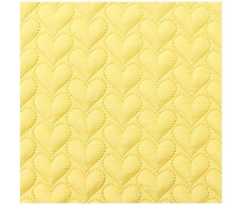 May include: Close-up of a yellow quilted fabric with a repeating heart pattern. The hearts are evenly spaced and outlined with stitching, creating a textured surface. The fabric has a soft, inviting appearance.
