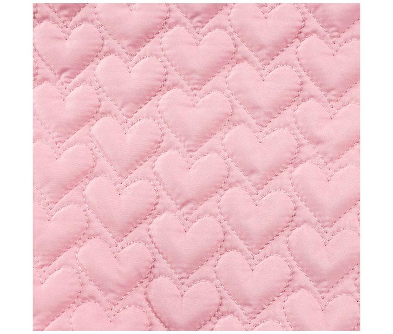 May include: Close-up of a pink quilted fabric with a repeating heart pattern. The hearts are evenly spaced and stitched onto the fabric, creating a textured surface. The color is a soft, pastel pink.