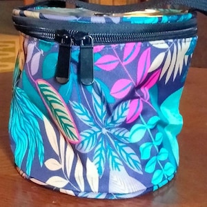 May include: A cylindrical, insulated lunch bag with a black zipper and strap. The bag features a vibrant tropical leaf pattern in shades of blue, teal, pink, and purple against a dark blue background. The bag is designed to keep food and drinks cool.