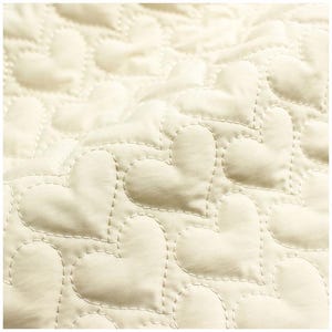 May include: Close-up of a cream-colored quilted fabric with a heart pattern. The hearts are stitched with a contrasting thread, creating a textured surface. The fabric has a soft, inviting appearance.