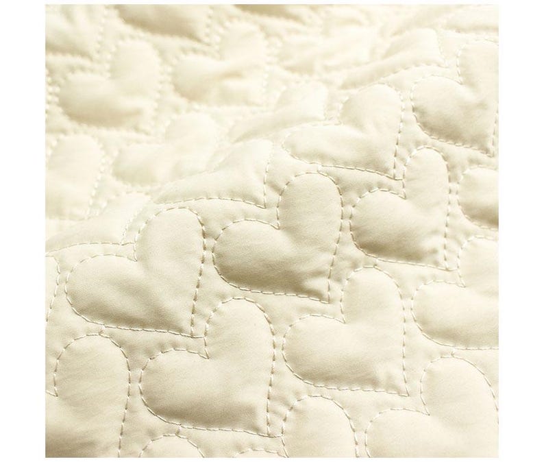 May include: Close-up of a cream-colored quilted fabric with a heart pattern. The hearts are stitched with a contrasting thread, creating a textured surface. The fabric has a soft, inviting appearance.