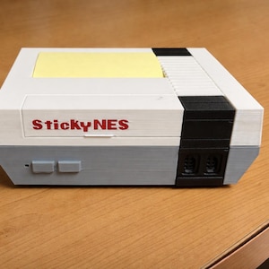Könnte beinhalten: Eine weiße, graue und schwarze Box, die einer klassischen Videospielkonsole ähnelt, mit dem Text "Sticky NES" in Rot. Die Oberseite hat einen gelben Haftnotizblock. Die Konsole hat zwei graue Tasten und zwei schwarze Anschlüsse.
