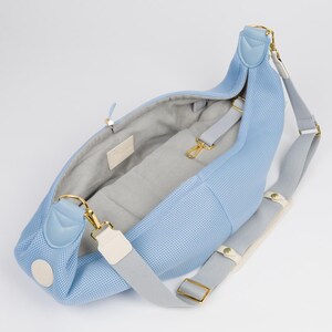 Peut inclure: Un sac de transport pour animaux de compagnie en maille bleu clair avec une sangle r&eacute;glable grise et blanche. Le sac a un fermoir en m&eacute;tal dor&eacute; et une doublure grise.