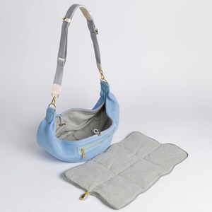 Peut inclure: Un sac &agrave; langer bleu clair avec une bandouli&egrave;re grise et blanche et un tapis &agrave; langer gris assorti. Le sac a une fermeture &eacute;clair et un tirette de fermeture &eacute;clair dor&eacute;e.