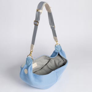 Peut inclure: Un sac de transport pour animaux de compagnie bleu clair avec une bandouli&egrave;re r&eacute;glable grise et blanche. Le sac est fait d'un mat&eacute;riau doux et respirant et poss&egrave;de un int&eacute;rieur rembourr&eacute;.