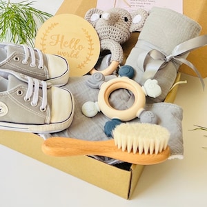 May include: A gift box filled with baby items, including a pair of gray baby shoes, a gray muslin swaddle blanket, a wooden teether with silicone rings, a crocheted elephant rattle, and a wooden baby brush.