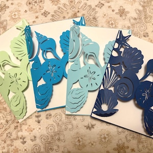 May include: Four white cards with laser-cut seashell designs in shades of green, blue, and navy. The cards feature intricate cutouts of starfish, shells, and sand dollars, creating a coastal theme.