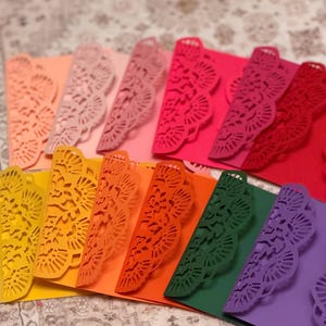 Peut inclure: Enveloppes en papier de couleurs vives, découpées au laser avec un bord décoratif. Les enveloppes sont dans des tons de rose, orange, jaune, vert et violet. Elles sont disposées en éventail, mettant en valeur leurs couleurs vives.