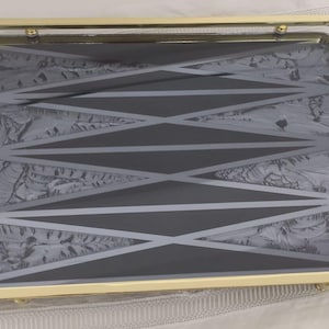 Puede incluir: Bandeja decorativa rectangular con marco y asas doradas. El interior de la bandeja presenta un patrón geométrico en tonos grises y blancos. Las asas tienen un diseño ornamentado y curvado.