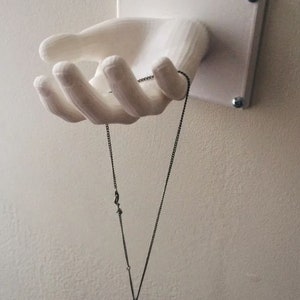 May include: A white 3D printed hand wall mount with a silver chain necklace hanging from it. The necklace has a small silver pendant.