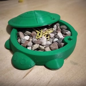May include: A green turtle-shaped container with a lid, filled with small rocks and paper clips. The container is made of plastic and has a smooth, glossy finish.