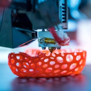 May include: A close-up of a 3D printer creating a red, lattice-like object. The printer nozzle is visible, extruding the plastic filament.