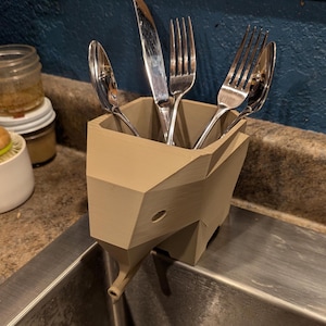 May include: A beige ceramic utensil holder shaped like an elephant with a spout. The holder is filled with a knife, two forks, and a spoon.