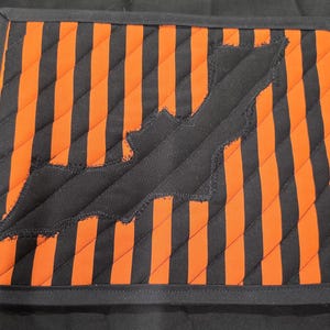 May include: A rectangular quilted placemat featuring a pattern of alternating black and orange diagonal stripes. A black fabric bat silhouette with raw, frayed edges is appliqued across the centre of the striped fabric. The entire piece is finished with a solid black fabric border.
