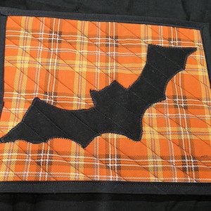 May include: A square quilted potholder featuring an orange, yellow, and brown tartan fabric pattern. A black fabric bat silhouette is appliqued in the centre of the square. The entire piece is finished with a solid black fabric binding border.