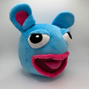 May include: A blue plush toy with large pink ears, a wide open mouth with a pink tongue sticking out, and large black eyes. The toy has a round body and a friendly expression.