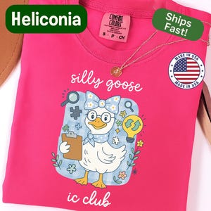May include: A hot pink t-shirt with a graphic of a goose wearing glasses and a bow tie, holding a clipboard. The text "silly goose" and "ic club" are printed on the shirt. A gold necklace is draped across the shirt.