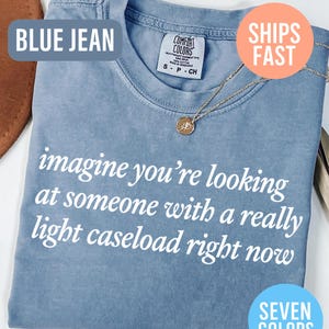 May include: Blue jean colored t-shirt with the text "imagine you're looking at someone with a really light caseload right now" in white. A gold necklace with a pendant is on the shirt. The shirt is labeled "Comfort Colors".