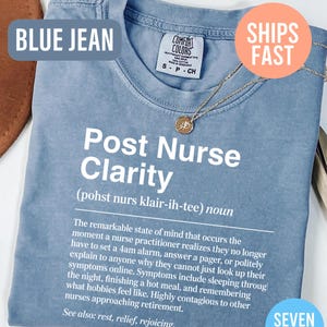May include: Blue jean colored t-shirt with the text "Post Nurse Clarity" and a definition. The shirt has a crew neck and short sleeves. A gold necklace with a pendant is draped on the shirt. The shirt is labeled "Comfort Colors".