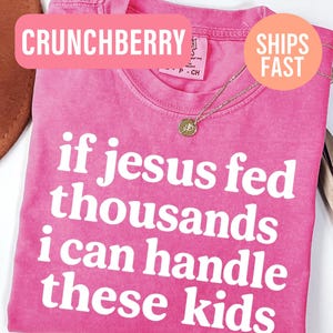 May include: A bright pink short-sleeved t-shirt featuring the white text "if jesus fed thousands i can handle these kids" printed on the front. The shirt has a crew neckline and a visible interior tag indicating size CH, with a gold-toned necklace.