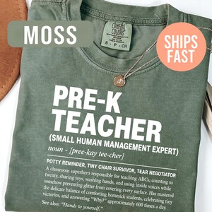 May include: A moss green crew neck t-shirt featuring white text that reads PRE-K TEACHER (SMALL HUMAN MANAGEMENT EXPERT) noun - [pree-kay tee-cher] POTTY REMINDER, TINY CHAIR SURVIVOR, TEAR NEGOTIATOR. A classroom superhero responsible for teaching ABCs.