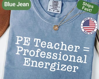 May include: A blue jean colored t-shirt with the text "PE Teacher = Professional Energizer" in white. The shirt has a crew neck and short sleeves. A gold necklace with a pendant is draped on the shirt. The shirt is labeled "Comfort Colors".