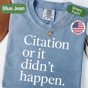 May include: A blue jean colored t-shirt with the text "Citation or it didn't happen" in white. The shirt has a crew neck and short sleeves. A gold necklace with a pendant is draped on the shirt. The shirt has a "Made in USA" tag.