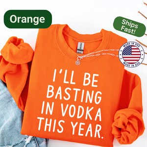 May include: Orange sweatshirt with the text "I'LL BE BASTING IN VODKA THIS YEAR" in white. The sweatshirt is folded on top of a pair of blue jeans. A silver necklace with a pendant is visible.