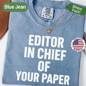 May include: A blue jean colored t-shirt with the text "EDITOR IN CHIEF OF YOUR PAPER" in white block letters. A gold necklace with a circular pendant rests on the shirt. The shirt has a Comfort Colors tag.