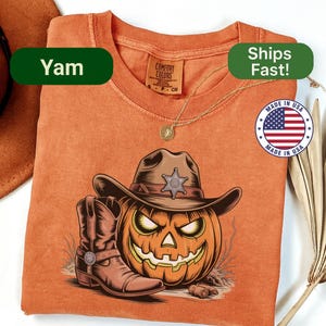 May include: Orange t-shirt featuring a Halloween design. The graphic includes a jack-o'-lantern wearing a cowboy hat and boots. The shirt has the text "Yam" and "Made in USA" on it.