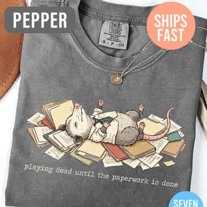May include: Pepper-colored t-shirt featuring a cartoon opossum lying on a pile of paperwork, wearing a white shirt and tie. The text "playing dead until the paperwork is done" is printed below the opossum. A silver necklace with a pendant is also visible.