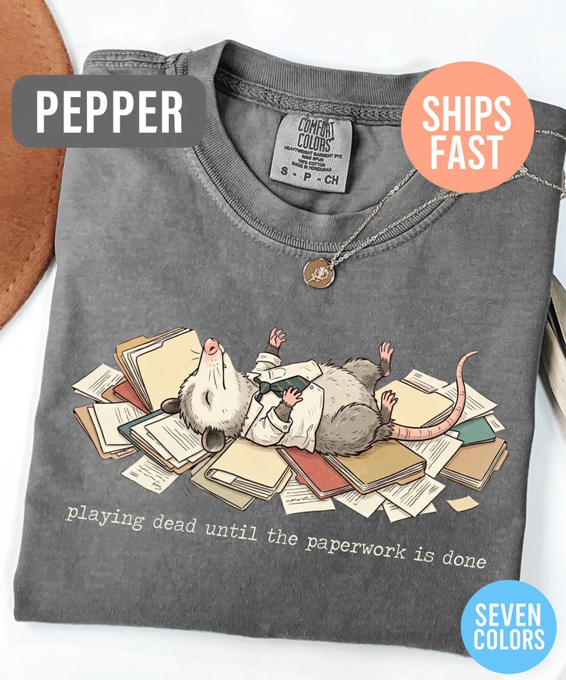 May include: Pepper-colored t-shirt featuring a cartoon opossum lying on a pile of paperwork, wearing a white shirt and tie. The text "playing dead until the paperwork is done" is printed below the opossum. A silver necklace with a pendant is also visible.