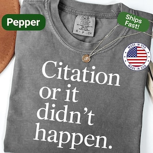 May include: A gray t-shirt with the text "Citation or it didn't happen" in white. The shirt has a crew neck and short sleeves. A silver necklace with a small pendant is draped across the shirt. The shirt is labeled "Pepper".